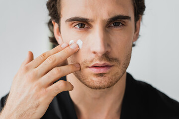 Young man looking at camera while applying cosmetic cream