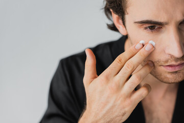 Cropped view of young man applying cream on face isolated on grey