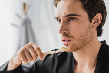 Young man in black robe holding wooden toothbrush with toothpaste in bathroom