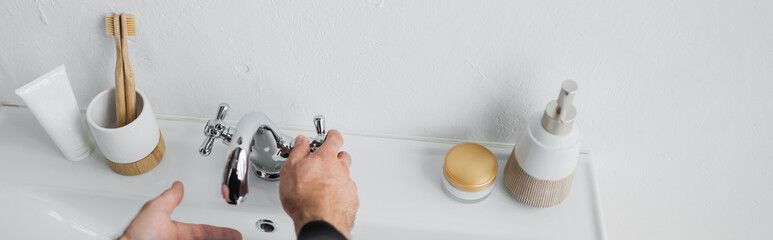 Cropped view of man tapping water near toiletries and sink in bathroom, banner