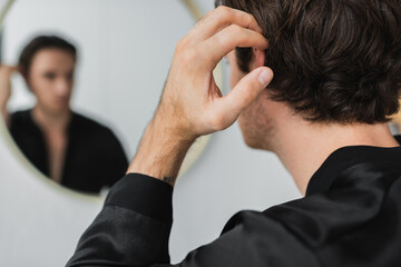 Young man in satin robe touching hair near blurred mirror at home