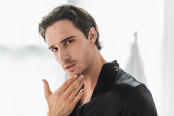 Young man in silk robe touching chin in bathroom