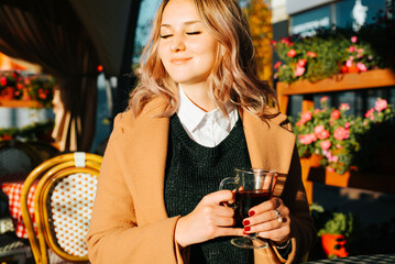 Portrait of pretty young blonde woman with curly hair and in coat with glass of mulled wine sitting in street cafe. Smiling lady with closed eyes relaxing enjoying break on terrace