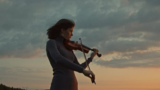 Woman musician is playing violin solo, performing outside, surrounded by forest, getting inspiration from nature, beautiful autumn sky is on the background, Slow motion.
