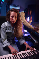 Fototapeta na wymiar Young female musician playing synthesizer in the recording studio with his colleague in the background