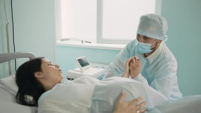Pregnant woman with painful contractions holding hand of her caring husband that sitting near at hospital ward. Young couple preparing for birth of their child.
