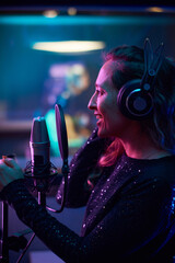 Naklejka premium Young woman in headphones singing to microphone while standing in the recording studio, she recording a new song