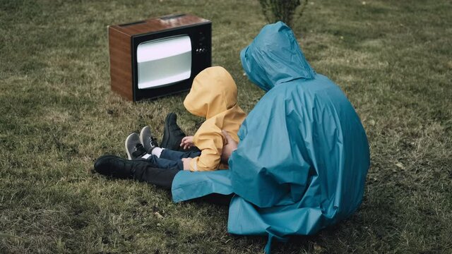 Woman with small child in raincoats with hood are sitting on grass and watching an old retro TV. TV is strobing. Concept of post apocalypse, horror, surrealism and mysticism. Dependence on television