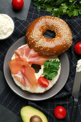 Delicious bagel with cream cheese, jamon, tomato and parsley on black table, flat lay