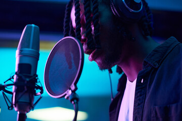Close-up of African man with dreadlocks singing to microphone during recording a new song in the music studio - obrazy, fototapety, plakaty