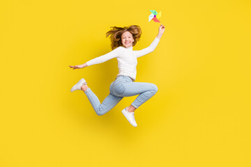 Full length photo of happy nice little girl jump up smile hold hand pinwheel isolated on yellow color background