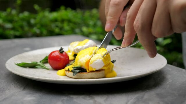 Healthy breakfast. Man eating delicious poached eggs at morning in green outdoor cafe. Male hands cuts egg benedict with liquid egg yolk inside, hollandaise sauce and spinach. Close up