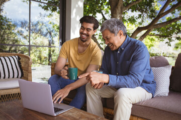 Smiling biracial adult son and senior father using laptop and drinking coffee in garden