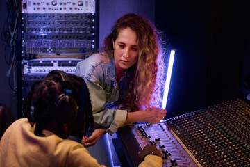 Young singer discussing new song together with her producer while he working on music keyboard in the studio