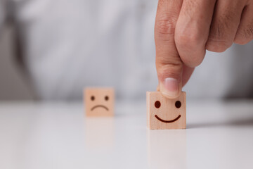 Customer hand in close-up On the wood cube, choose a happy face and a blurred sad face symbol, Service rating, and satisfaction idea.