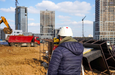 Photo of a construction worker in a white helmet who looks into the level at the construction site of multi-storey buildings with a red truck on the street in the city