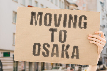 The phrase " Moving to Osaka " drawn on a carton banner in men's hand. Human holds a cardboard with an inscription. Big city. Urban. Opportunity. Immigration