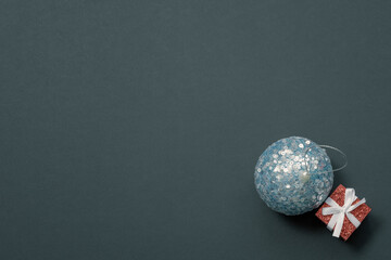 A shiny blue Christmas ball in sequins lies next to a miniature red box on a dark greenish background. Christmas card.