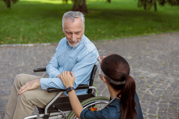 Obraz premium Senior aged man, recovering patient in wheelchair looking and holding hand of nurse wearing face shield and mask, supporting him