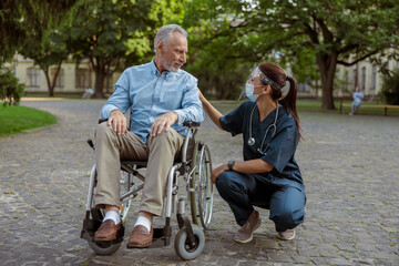 Full length shot of caring nurse wearing face shield and mask talking to mature man, recovering patient in wheelchair on a walk in the park near hospital
