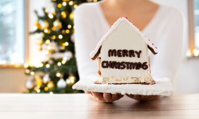winter holidays, baking and cooking concept - close up of woman with gingerbread house at home over christmas tree lights on background