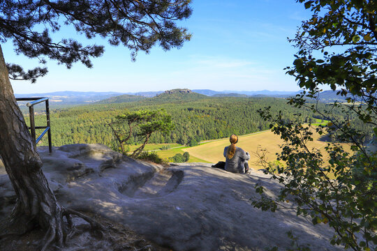 A woman is sitting on mountain Pfaffenstein with view at mountain Gohrischstein, Saxon Switzerland - Germany