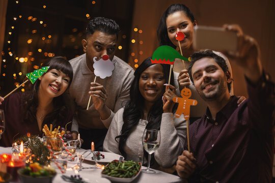 Holidays, People And Celebration Concept - Multiethnic Group Of Happy Friends With Party Props Having Christmas Dinner At Home And Taking Selfie With Smartphone