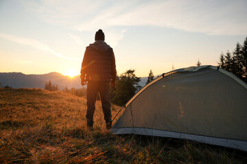 Man near camping tent in mountains at sunset, back view - obrazy, fototapety, plakaty