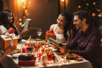 holidays, party and celebration concept - multiethnic group of happy friends having christmas dinner and giving presents at home