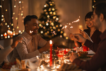 holidays and celebration concept - multiethnic group of happy friends with sparklers having christmas dinner at home