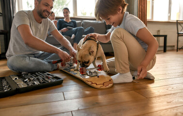 Smiling father and son fix skateboard play with dog