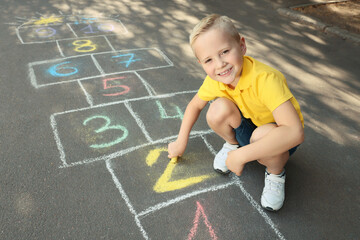 Little boy drawing hopscotch with chalk on asphalt outdoors. Happy childhood
