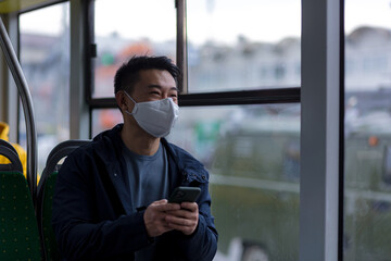 An Asian passenger in a protective medical mask on his face anxiously writes and reads news from a mobile phone, a man travels around the city by public transport bus