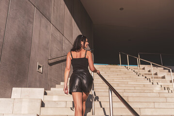 Dark-haired woman posing in a black dress on the concrete stairs. The woman is wearing black sunglasses. It's a beautiful sunny day.