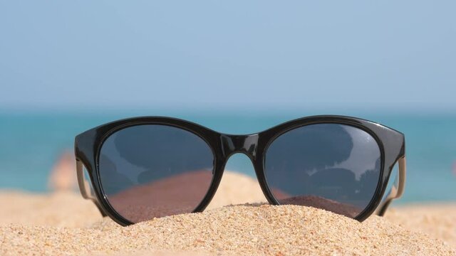 Closeup of black protective sunglasses on sandy beach at tropical seaside on warm sunny day. Summer vacation concept.