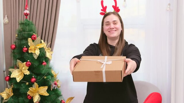 Young woman wearing deer horns holding the present box and covering her face with it, then making a giving gesture of the gift standing near the New Year tree.