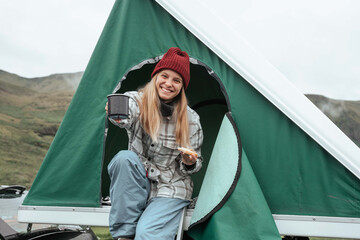 Caucasian girl stretching cup of hot tea to the camera while sitting at the tent