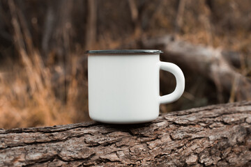 White blank metal mug mock up standing on tree outdoors, autumn forest behind. Enamelled travel cup...