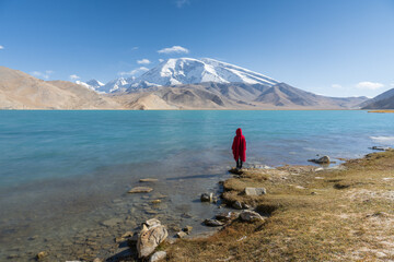 A woman in red cloak stands by the lake and looks at the Muztagh Ata peak.
