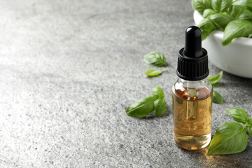 Glass bottle of basil essential oil and leaves on grey stone table. Space for text