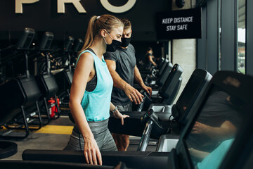 Fototapeta na wymiar Young fit woman and man running on treadmill in modern fitness gym. They keeping distance and wearing protective face masks. Coronavirus world pandemic and sport theme.