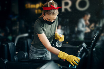 Young female worker disinfecting cleaning and weeping expensive fitness gym equipment with alcohol sprayer and cloth. Coronavirus global world pandemic and health protection safety measures.