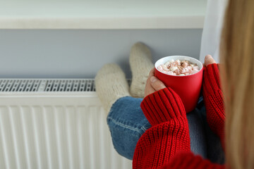 Woman with cup of cocoa warms her feet by the radiator