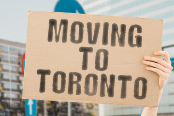The phrase " Moving to Toronto " drawn on a carton banner in men's hand. Human holds a cardboard with an inscription. Big city. Urban. Opportunity. Immigration
