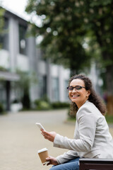 Fototapeta premium african american businesswoman with takeaway drink and mobile phone smiling at camera while sitting outdoors