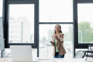 african american businesswoman with smartphone pointing at blurred laptop during video conference in office