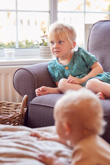 Young Girl Sitting On Sofa In Lounge At Home Watching TV As Baby Brother Plays In Foreground