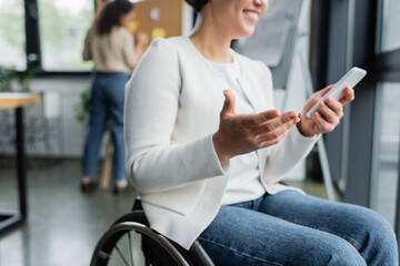 cropped view of cheerful businesswoman in wheelchair gesturing while using mobile phone near...