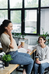 cheerful african american businesswoman adjusting earphone while sitting on desk near colleague in wheelchair