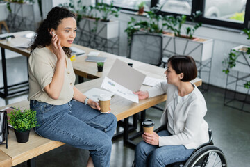 businesswoman in wheelchair looking at infographics near african american colleague sitting on desk during coffee break